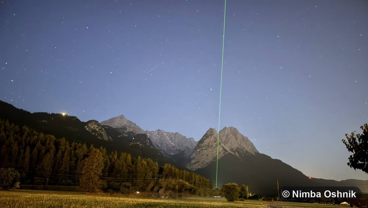 Green laser beam rising over moonlit mountains into a starry sky